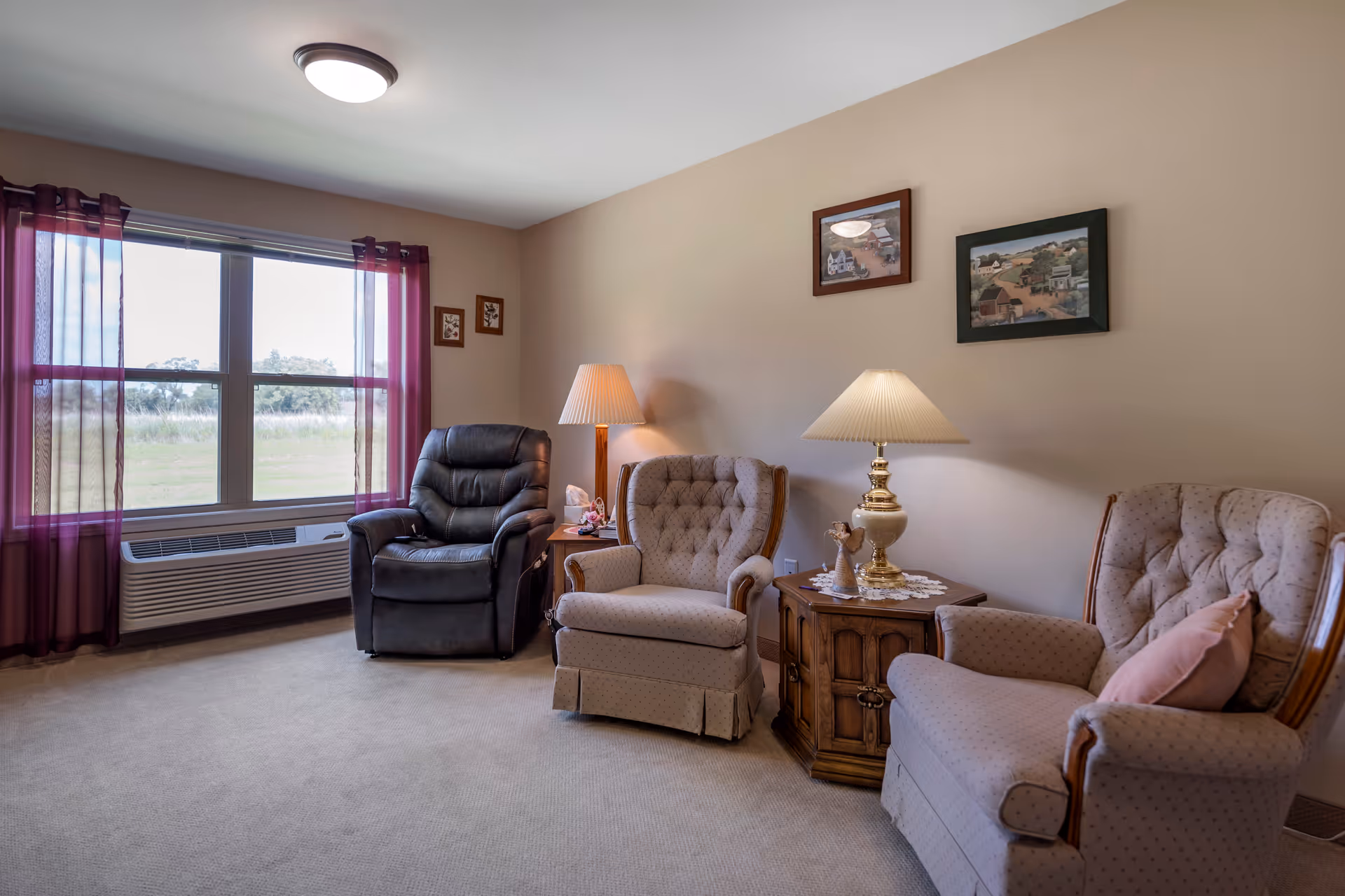 A cozy living room with three armchairs, two beige and one black recliner, arranged around a wooden side table with a lamp and decorative items. The room has beige walls with framed artwork and a large window with sheer purple curtains letting in natural light.