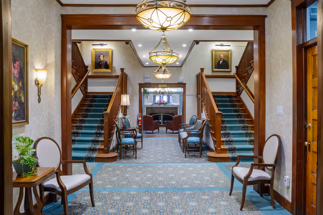 Interior view of a traditional common area in a senior living facility featuring a symmetrical double staircase with green carpet runners, wooden railings, and framed portraits above each staircase. The room has patterned carpet flooring, several upholstered chairs along the walls, a small table with a plant, and elegant hanging light fixtures. In the background, there is a seating area with armchairs near a fireplace.