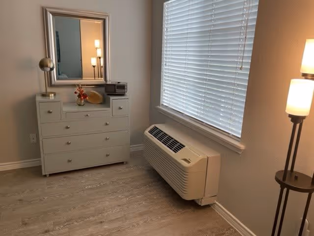 A corner of a room featuring a white dresser with multiple drawers, a mirror above it, and decorative items including a globe and a small vase with flowers. Next to the dresser is a window with closed blinds and a wall-mounted air conditioning unit below it. A floor lamp with two lit bulbs stands to the right of the window. The room has light-colored wooden flooring and neutral-colored walls.