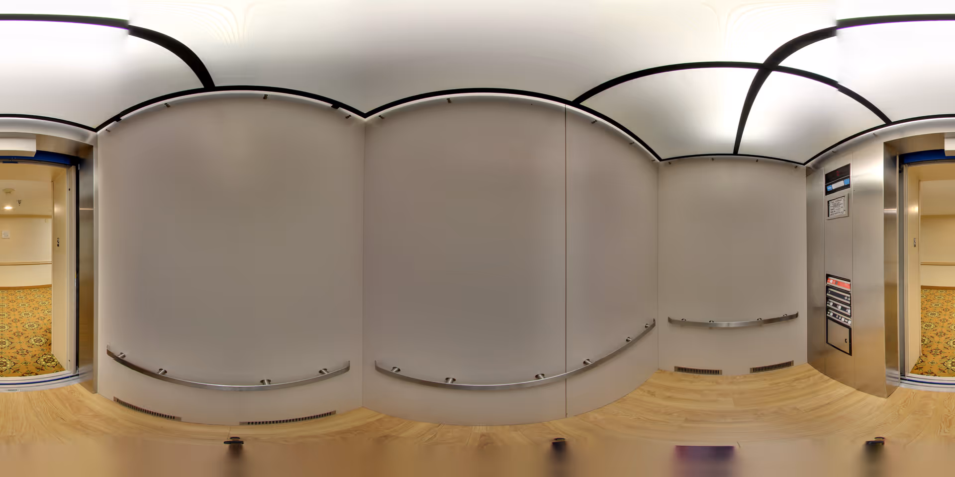 Interior of an elevator with light-colored walls, wooden floor, and metal handrails on three sides. The elevator doors are open, showing a hallway with patterned carpet outside. The ceiling has a unique curved design with bright lighting.