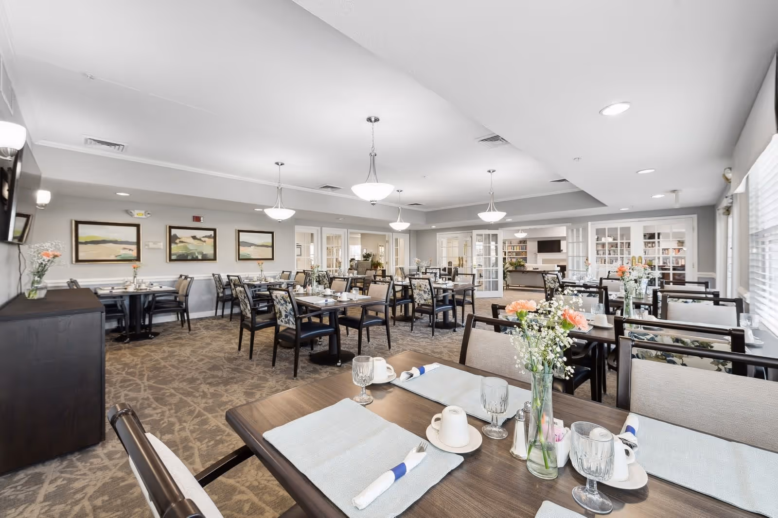 Bright, spacious dining room with multiple set tables and chairs, pendant lights, floral centerpieces, and large windows.