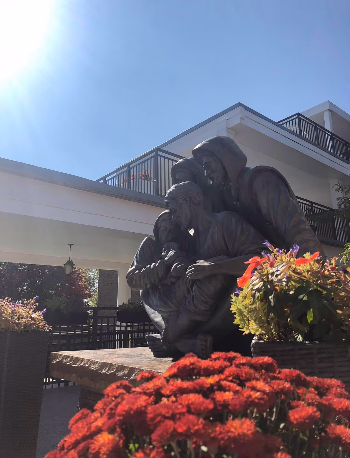 Bronze sculpture of three seated figures surrounded by flower planters in front of a multi-story building under a bright sky.