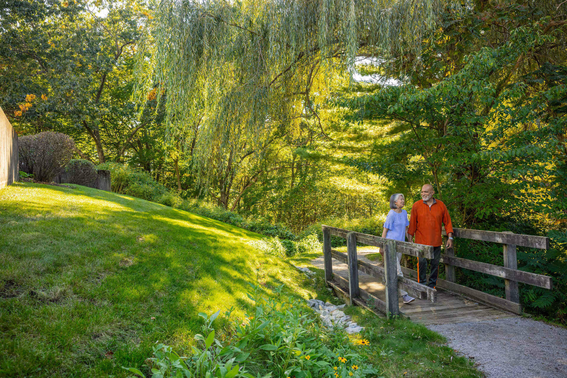 An elderly couple walking hand in hand across a small wooden bridge in a lush, green outdoor garden area with trees and grass surrounding them.