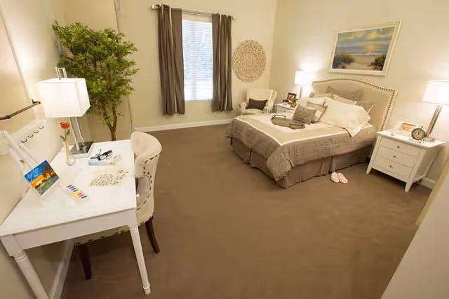 A cozy bedroom with a neatly made bed featuring beige and white bedding, a white nightstand with a lamp and clock, a white desk with a chair and desk lamp, a potted plant, and a window with brown curtains letting in natural light.