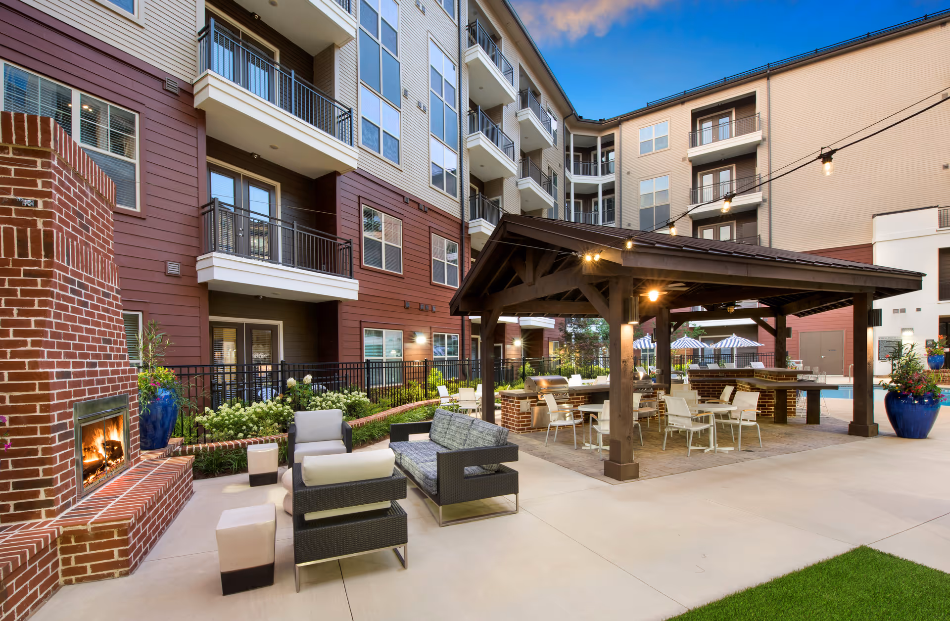 Outdoor patio area at Overture Buckhead South featuring a brick fireplace with a fire burning, modern outdoor seating with cushioned chairs and a sofa, a covered pavilion with tables and chairs, and string lights overhead. The multi-story building with balconies surrounds the patio area under a clear blue sky.