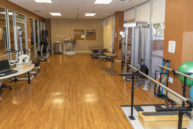 A spacious physical therapy or rehabilitation room with wooden flooring, exercise equipment including parallel bars, a stationary bike, therapy tables, and various exercise tools. Large windows on one side allow natural light to enter the room.