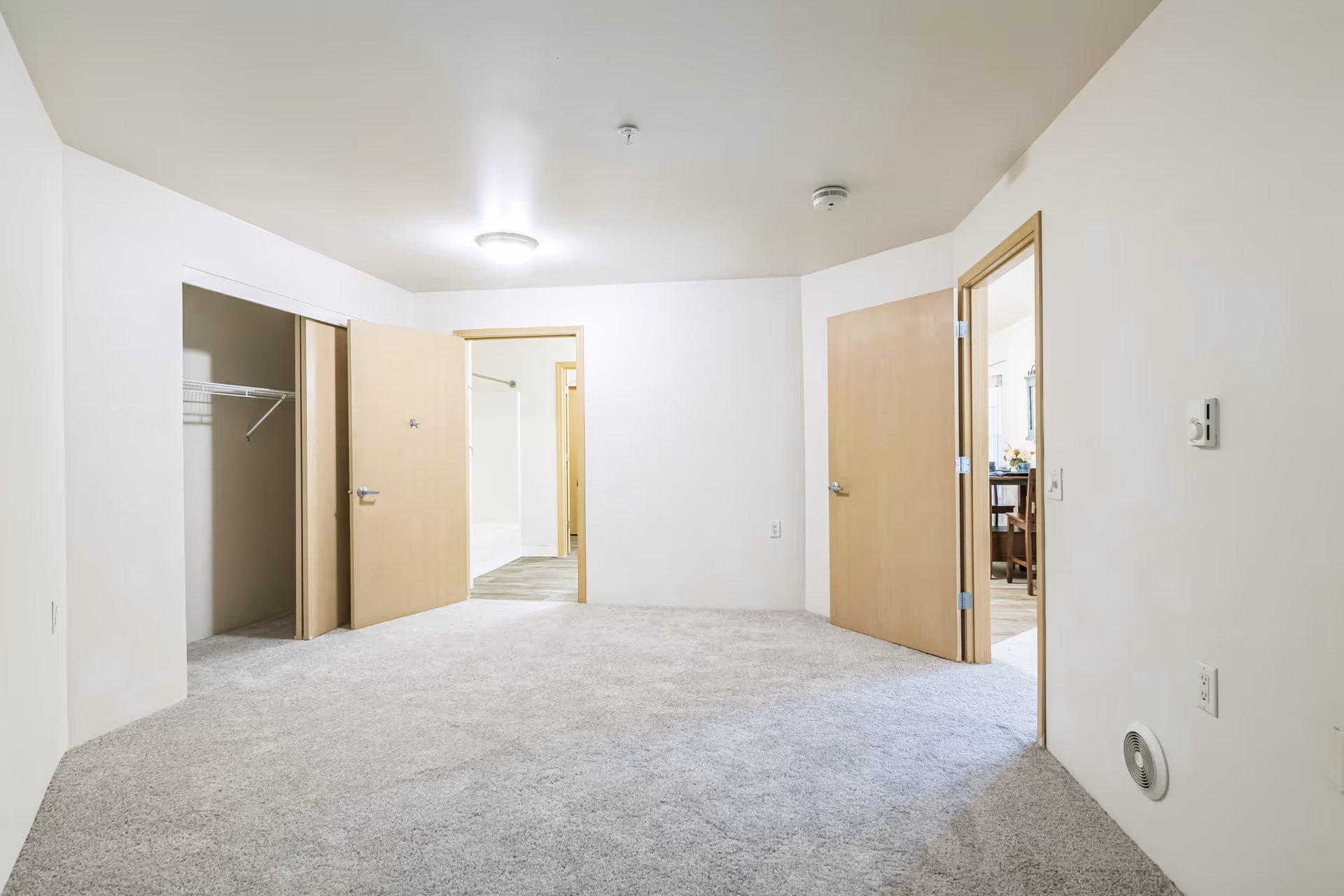 Empty room with light beige carpet and white walls featuring three open wooden doors. One door reveals a closet with a wire shelf, another door leads to a bathroom with a bathtub and shower curtain, and the third door opens to a dining area with a table and chairs. The ceiling has a round light fixture and a smoke detector.