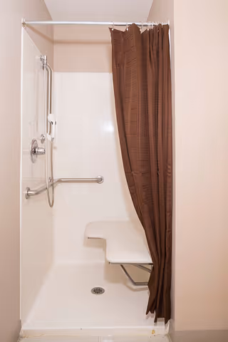 A small shower stall with a white fold-down seat, a handheld showerhead attached to a vertical grab bar, and a brown shower curtain partially drawn to the side.