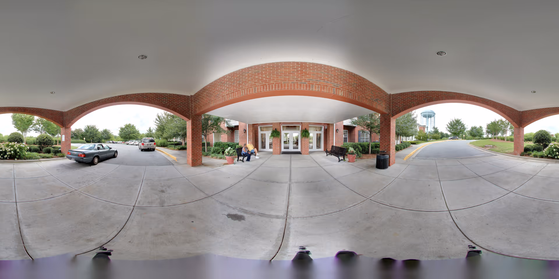 Covered entrance area of a brick building with three large arches supported by brick columns. Two people are sitting on a bench near the entrance doors. There are potted plants and a trash can near the entrance. Cars are parked on the driveway, and trees and greenery surround the area. A water tower is visible in the background.