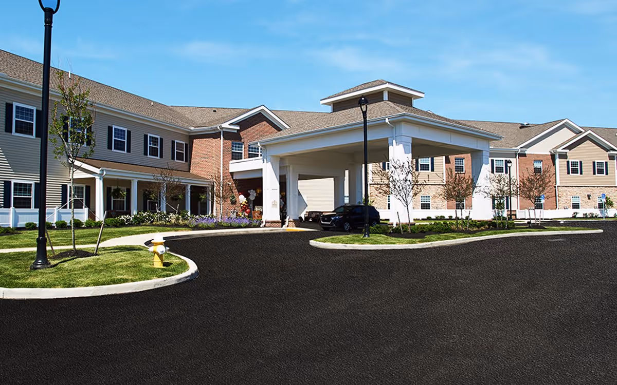 Exterior view of Grove City Senior Living facility showing a large two-story building with beige and brick facade, multiple windows, a covered entrance with white columns, a driveway, landscaped grass areas, small trees, and a fire hydrant under a clear blue sky.