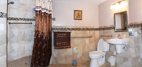 A bathroom with beige tiled walls and floor, featuring a walk-in shower with a brown and white shower curtain, a white toilet with the lid open, a wall-mounted white sink with a mirror above it, a towel rack with a brown towel, a toilet paper holder, and a small framed picture on the wall.
