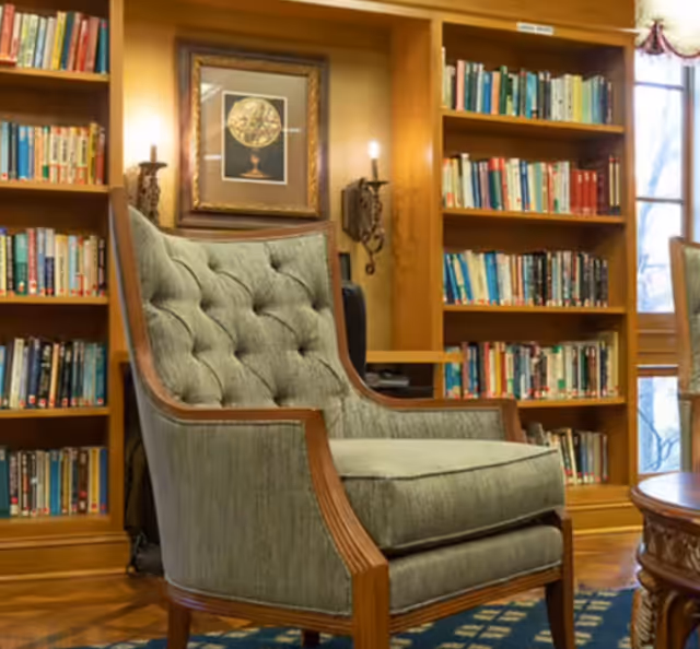A cozy reading area featuring a tufted armchair with wooden frame placed in front of wooden bookshelves filled with books. The background includes a framed picture of a globe and wall-mounted candle sconces, with a window partially visible on the right side.