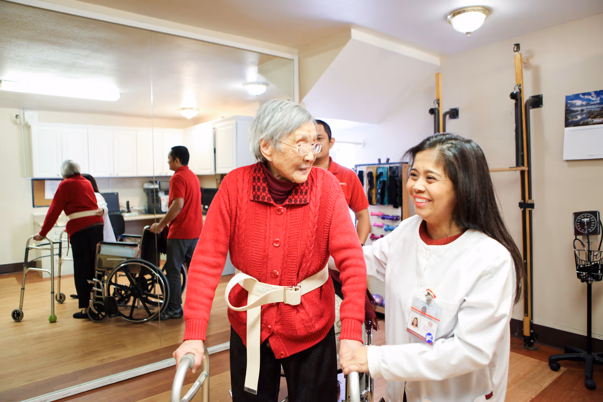 An elderly woman using a walker is assisted by a smiling healthcare worker in a rehabilitation or therapy room. In the background, another elderly person with a walker and a wheelchair are visible, along with a man in a red shirt. The room has wooden floors, white cabinets, and various therapy equipment.