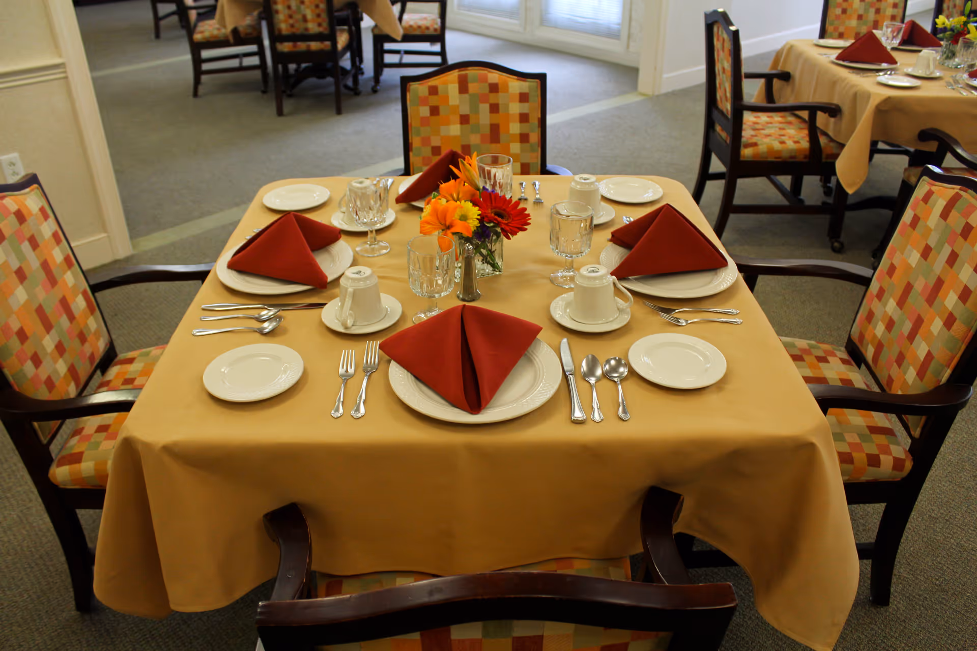 A dining table set for four with a beige tablecloth, red folded napkins, white plates, cups, glasses, and silverware. A small vase with colorful flowers is placed in the center of the table. The chairs around the table have a patterned upholstery with shades of orange, yellow, and beige. Additional similarly set tables are visible in the background.
