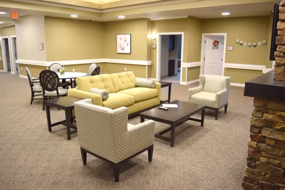 A cozy living room area in a senior living facility with a yellow sofa, two patterned armchairs, a dark wooden coffee table, and side tables. In the background, there is a round dining table with four chairs, beige carpet, light green walls, and a stone fireplace on the right side.