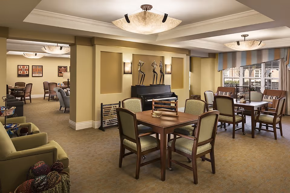 Spacious, well-lit communal lounge with multiple tables and chairs, a piano, and comfortable seating near large windows.