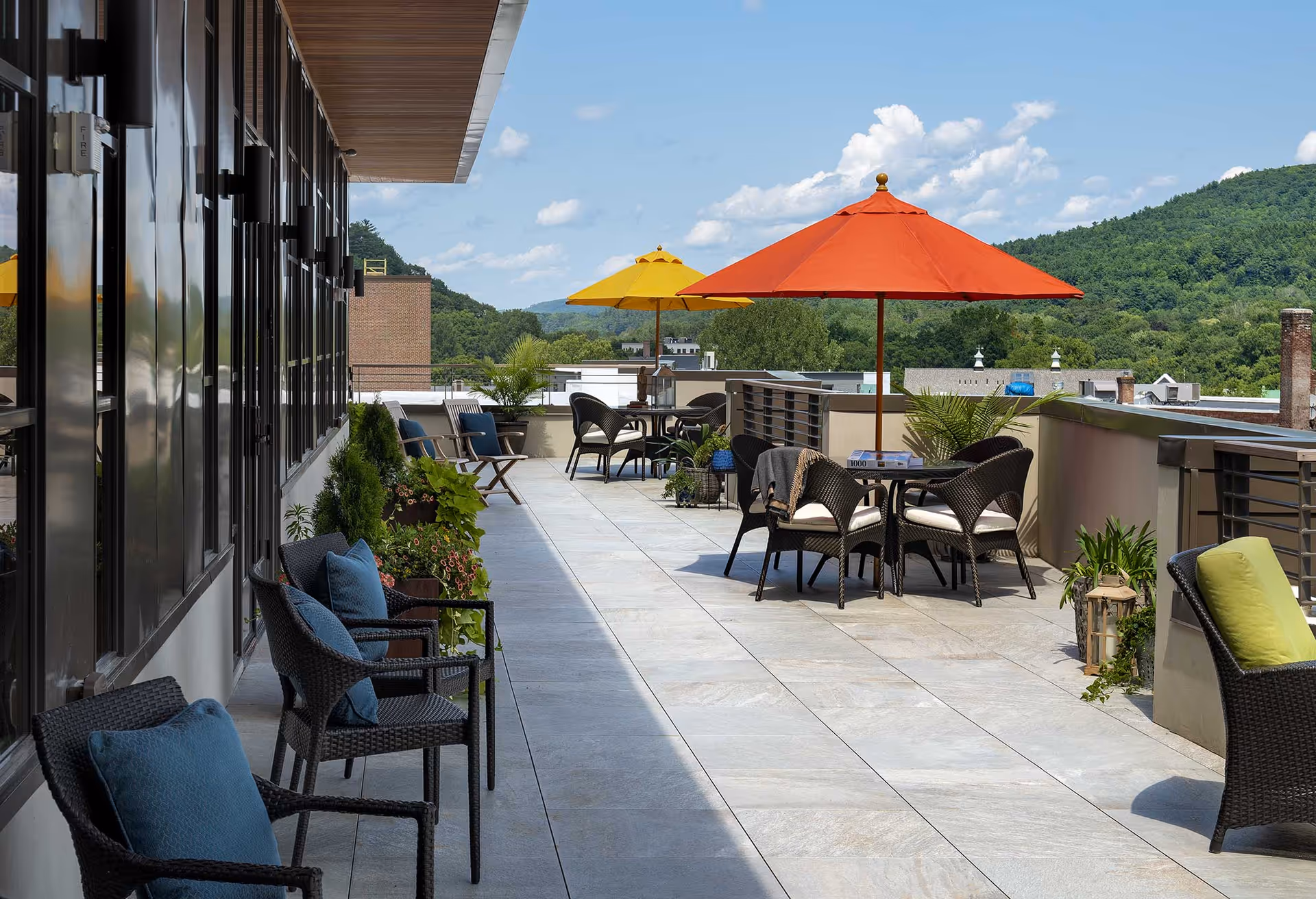Rooftop terrace with wicker chairs and tables, colorful umbrellas, potted plants and mountain views.