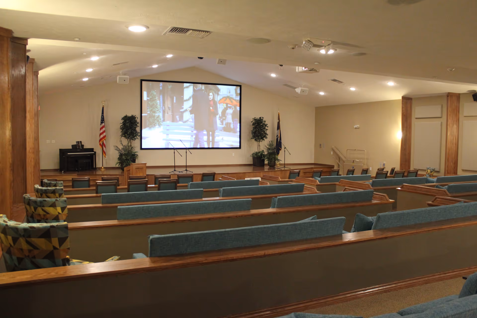 Interior view of a large room with rows of wooden pew-style seating with blue cushions facing a stage area. On the stage, there is a large screen displaying an image, two microphones on stands, a piano, and two potted plants. An American flag and another flag are positioned on either side of the stage. The room has a beige color scheme with wooden columns and ceiling lights.