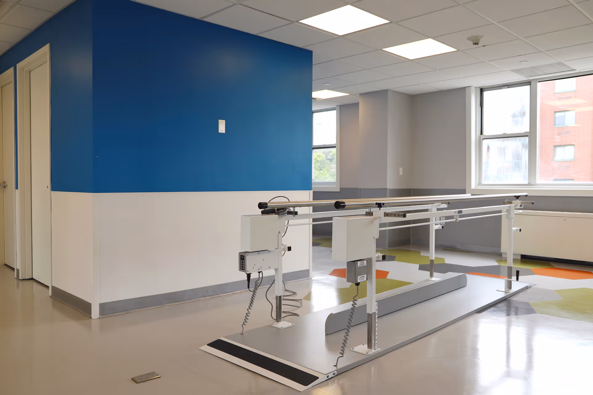 A rehabilitation room with parallel bars for physical therapy, featuring a blue and white wall, large windows letting in natural light, and a colorful patterned floor.