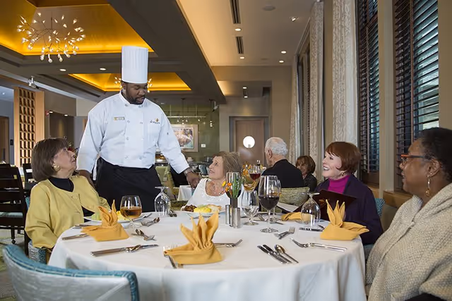 A chef in a white uniform and tall hat interacts with four elderly women seated around a round dining table set with yellow napkins, silverware, and glasses of wine in a warmly lit dining room with large windows and modern decor.