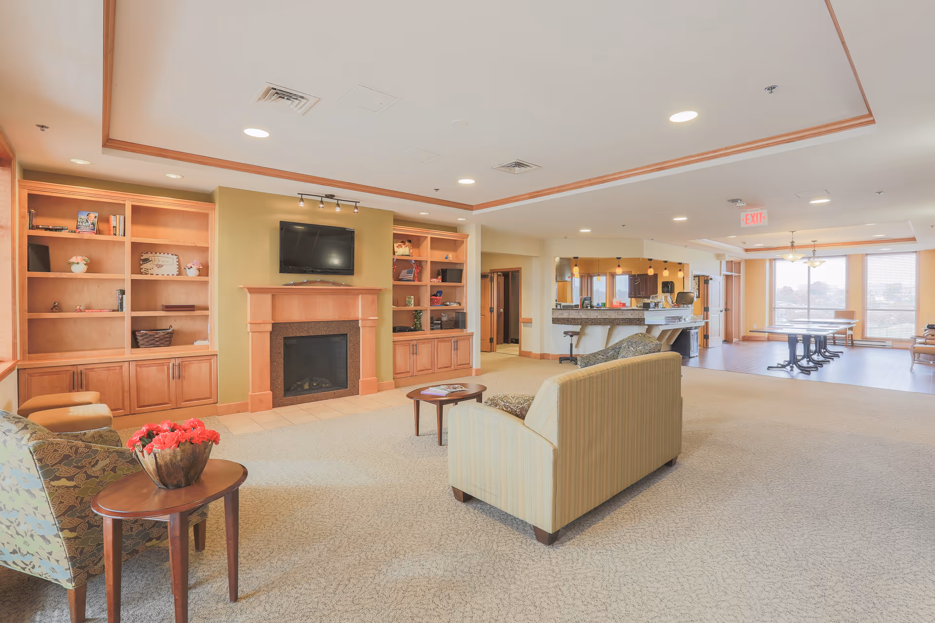 A spacious and well-lit common area in a senior living facility featuring a fireplace with a mounted TV above it, built-in wooden shelves on either side, comfortable seating including a striped sofa and patterned armchair, a small wooden table with a flower arrangement, and a dining area with tables and chairs near large windows letting in natural light.