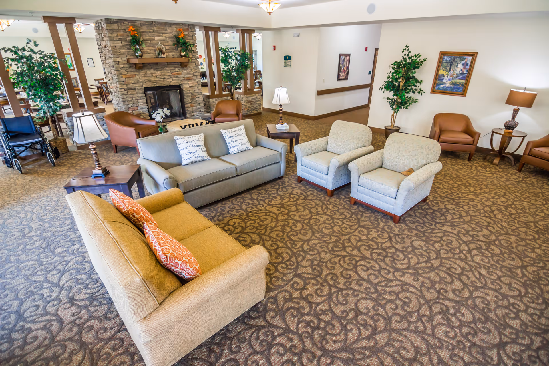 A spacious senior living facility common area with patterned carpet, multiple seating options including sofas, armchairs, and side tables with lamps. A stone fireplace is centrally located with decorative plants and artwork on the walls. A wheelchair is visible near the left side of the room.