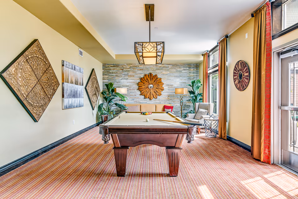 A well-lit recreational room with a pool table in the center, surrounded by decorative wall art, potted plants, a sofa, an armchair, and large windows with curtains allowing natural light to enter.