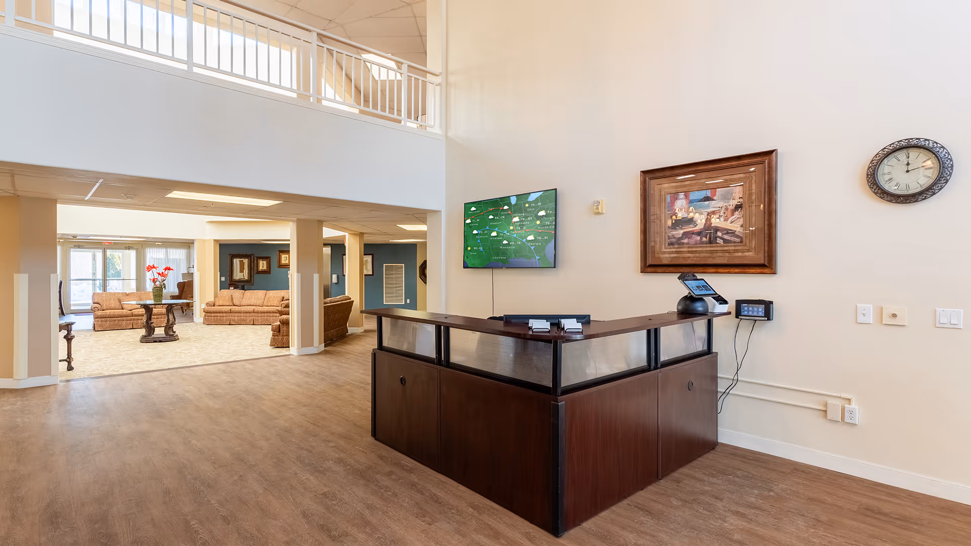 Interior view of a senior living facility lobby area with a dark wood reception desk in the foreground. Behind the desk, a wall-mounted TV displays a map, and a framed painting and clock are on the wall. In the background, there is a seating area with multiple brown sofas and a round table with a vase of red flowers. The space has high ceilings and large windows allowing natural light.