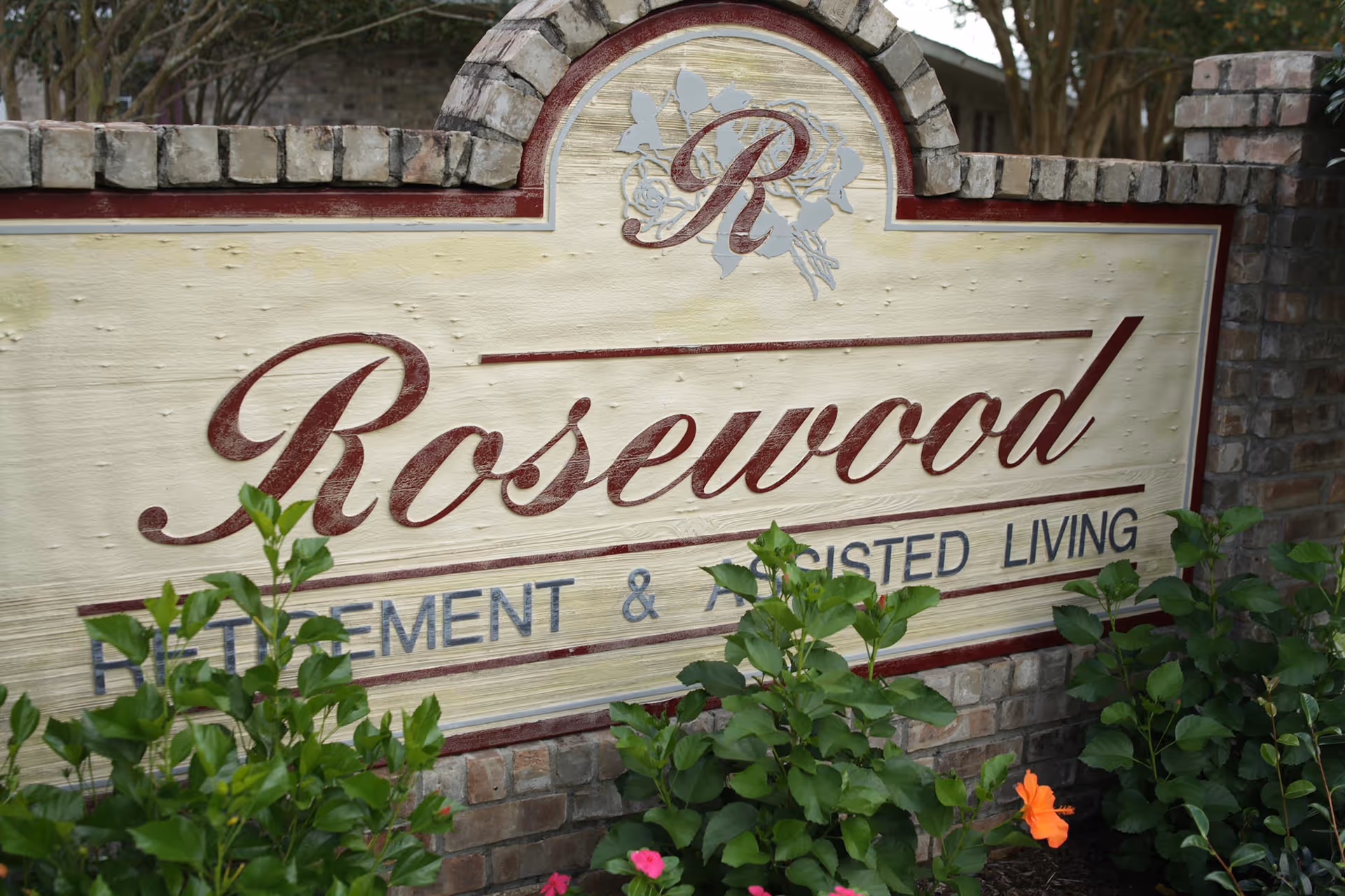 A stone and brick sign for Rosewood Retirement & Assisted Living surrounded by green plants and flowers.