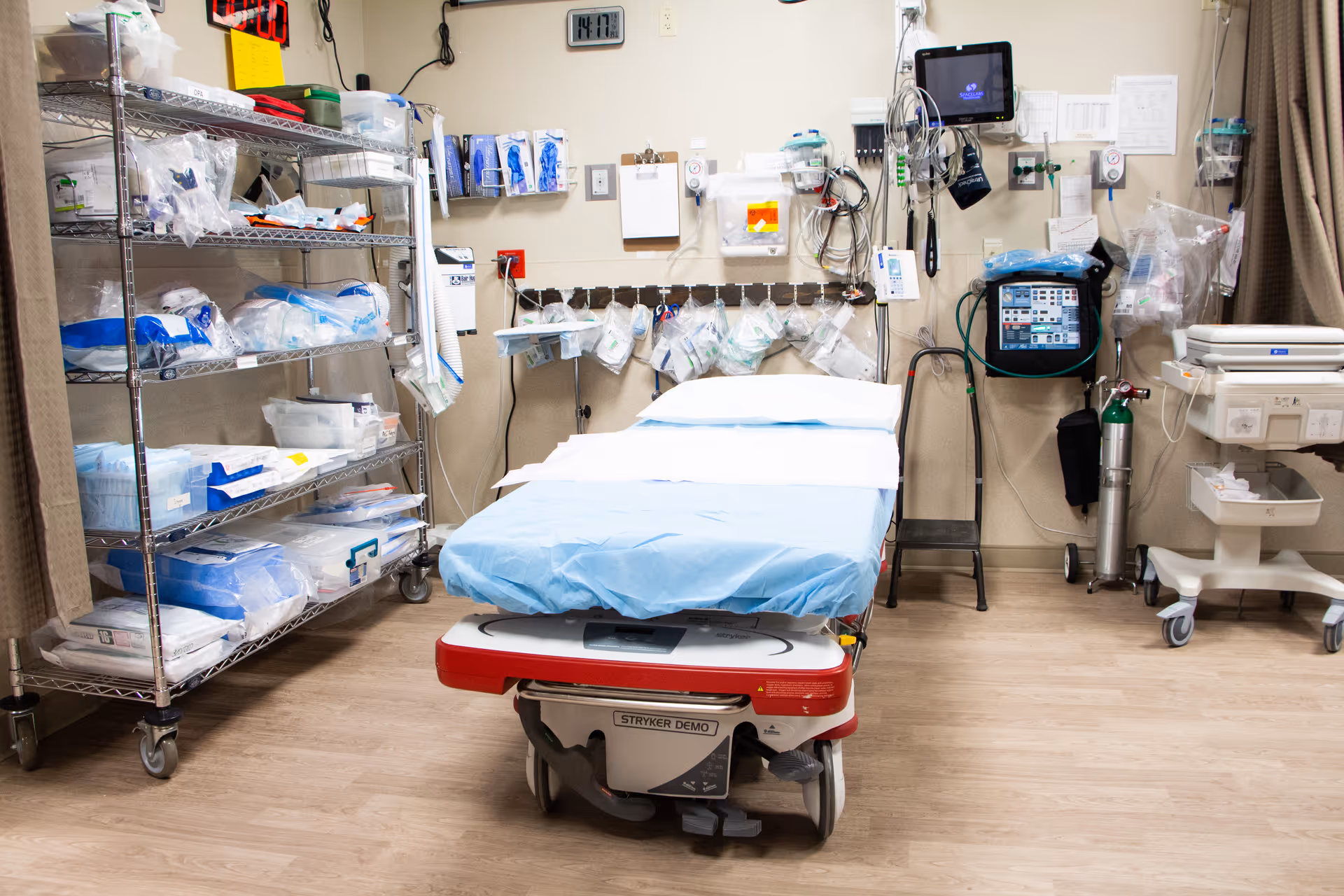 A medical examination or treatment room with a hospital bed covered in blue sheets in the center. The room has medical equipment mounted on the walls, including monitors, oxygen tanks, and various medical supplies organized on metal shelves and hanging hooks. The floor is wood-patterned, and the walls are beige.