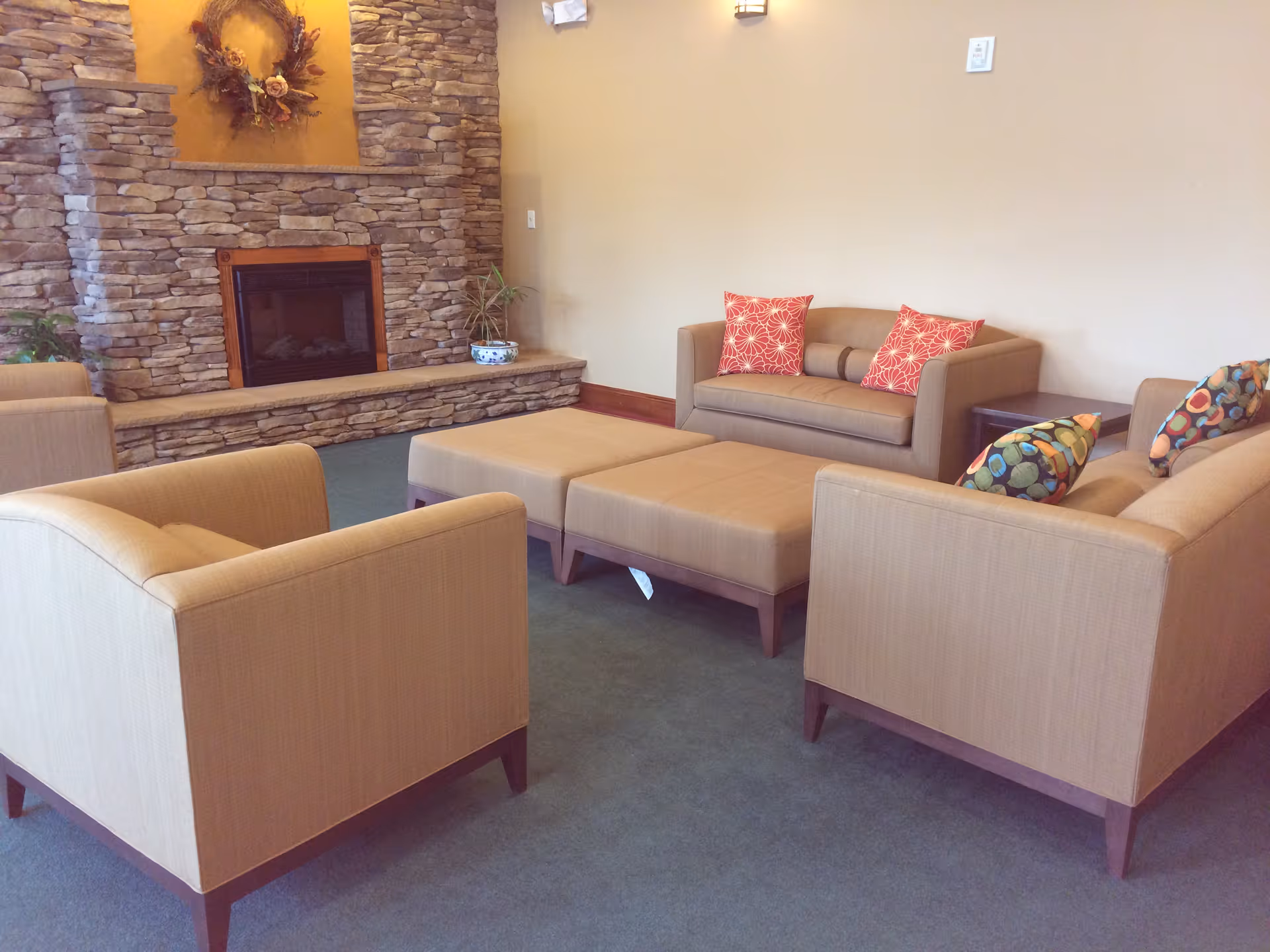 A cozy living room area with beige armchairs and a loveseat adorned with colorful patterned pillows. In the background, there is a stone fireplace with a decorative wreath above it and a small potted plant on the hearth.