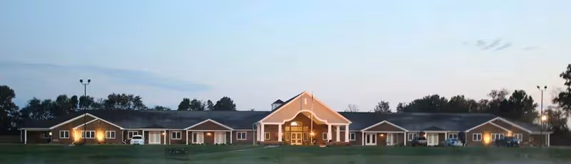 Front exterior of a long single-story senior living building with a central portico entrance, lit windows, and a lawn at dusk.