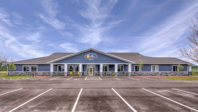 Front exterior of a single-story BeeHive Homes building with a wide empty parking lot under a blue sky.