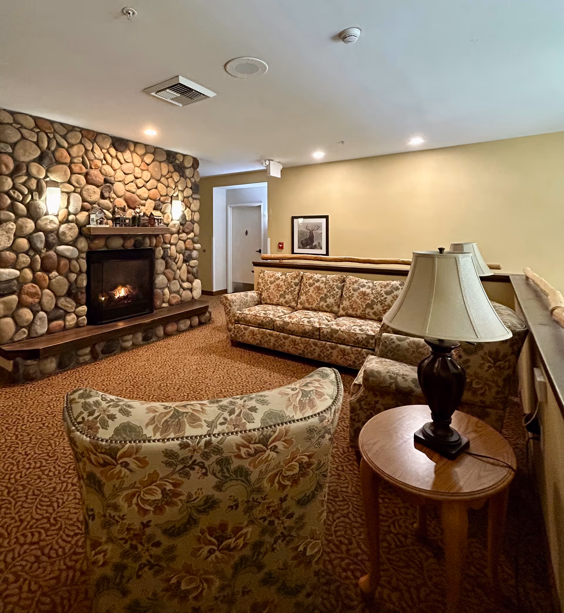 Cozy senior living room with a stone fireplace, a floral patterned sofa and armchair, a wooden side table with a lamp, beige walls, and carpeted floor.