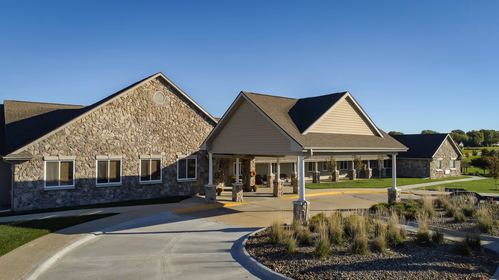 Exterior view of a single-story assisted living facility with stone and siding facade, a covered entrance supported by columns, and landscaped grounds with grass and ornamental grasses under a clear blue sky.