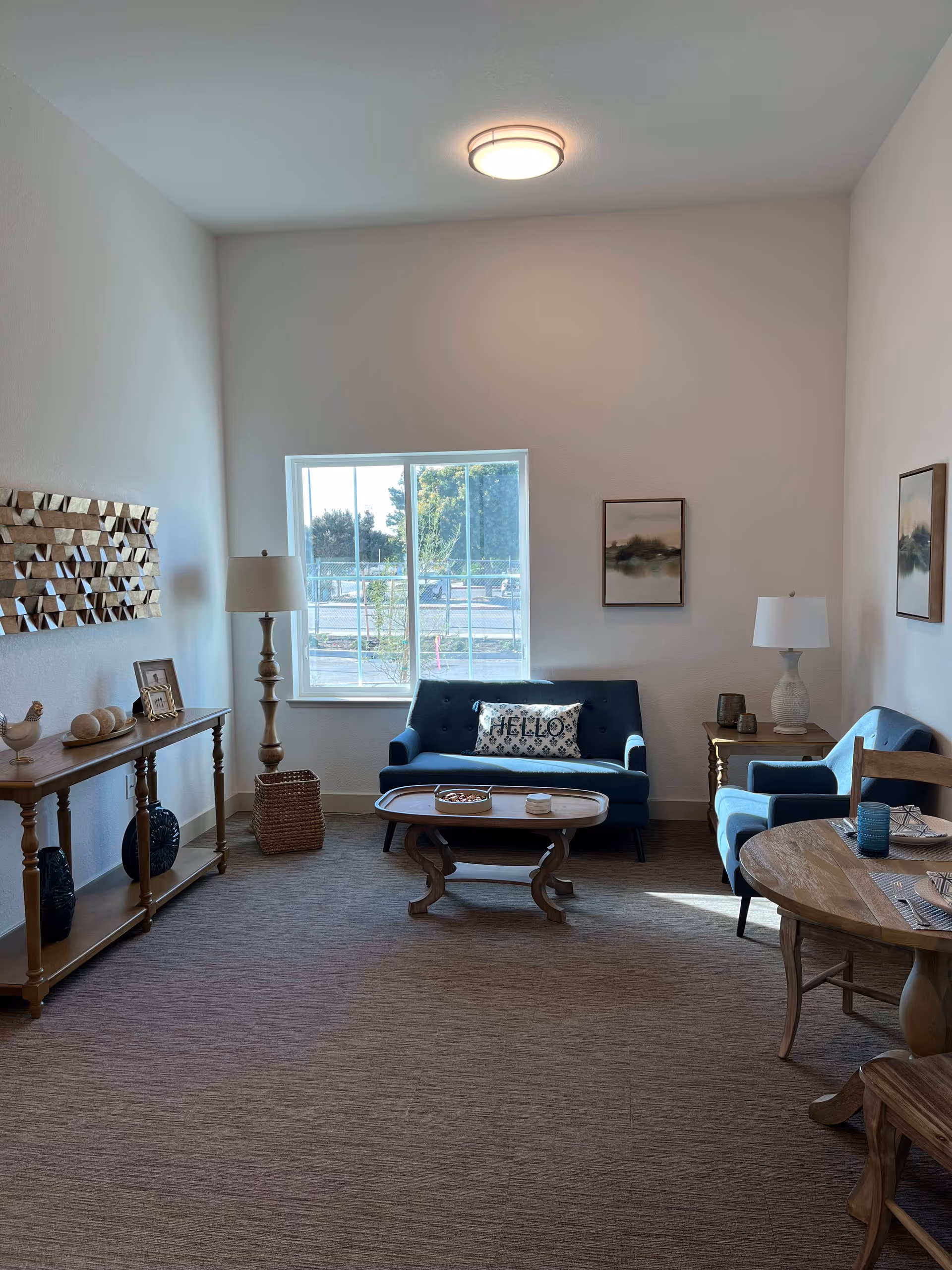 A cozy living room with a blue loveseat and matching armchair, a wooden coffee table, and a round wooden dining table with two chairs. The room has a large window letting in natural light, two table lamps, wall art, and a wooden console table with decorative items.