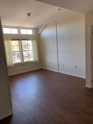 Empty room with wooden flooring, white walls, and a large window with sheer curtains allowing natural light to enter.