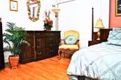A cozy bedroom in a senior living facility featuring a bed, dresser, chair, and plants.
