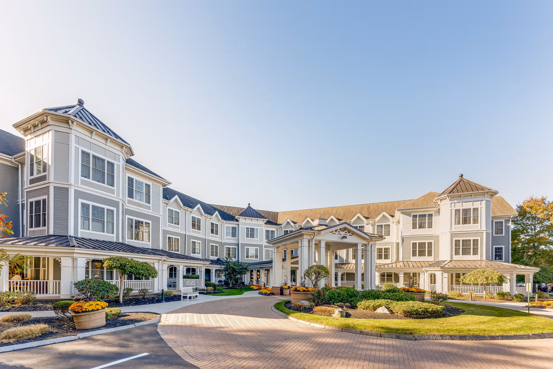 Exterior view of a large, three-story senior living facility building with multiple windows, a covered entrance with columns, landscaped gardens, and a clear blue sky.