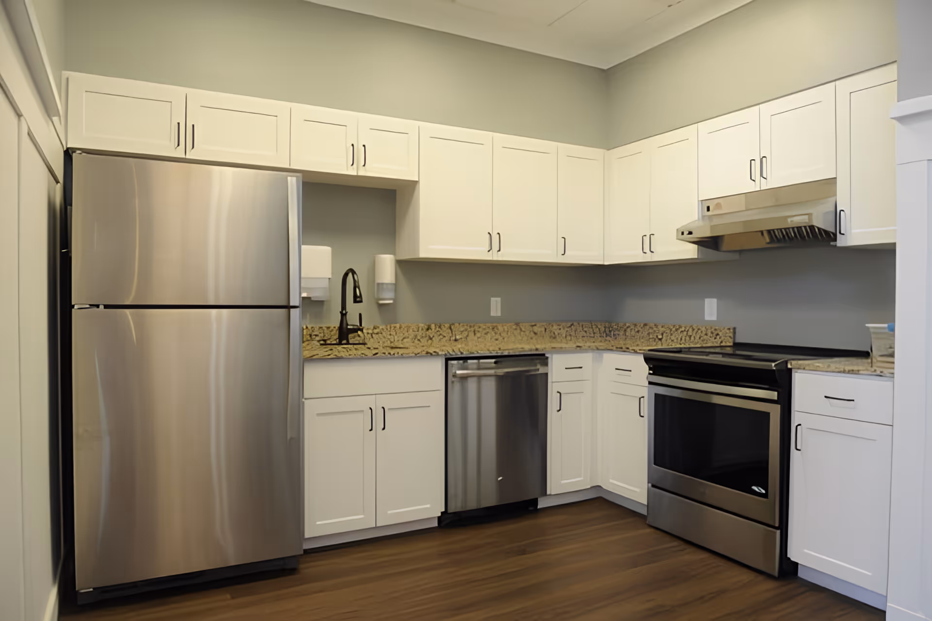 Modern kitchen with stainless steel refrigerator, dishwasher, and oven. White cabinets with black handles, granite countertops, and a black faucet. The floor is wooden, and the walls are painted light gray.