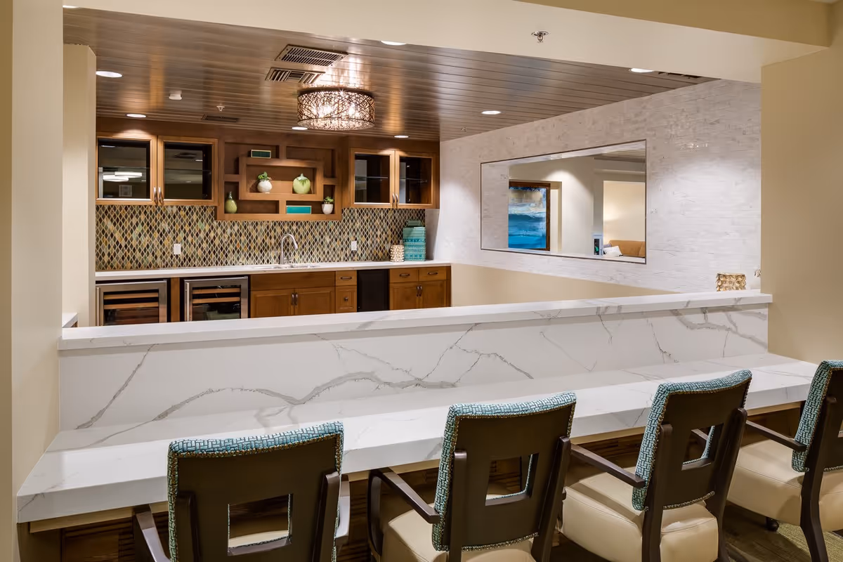 A modern communal bar/kitchen area with a marble countertop, four upholstered bar stools, wooden cabinets, and a mosaic tile backsplash.