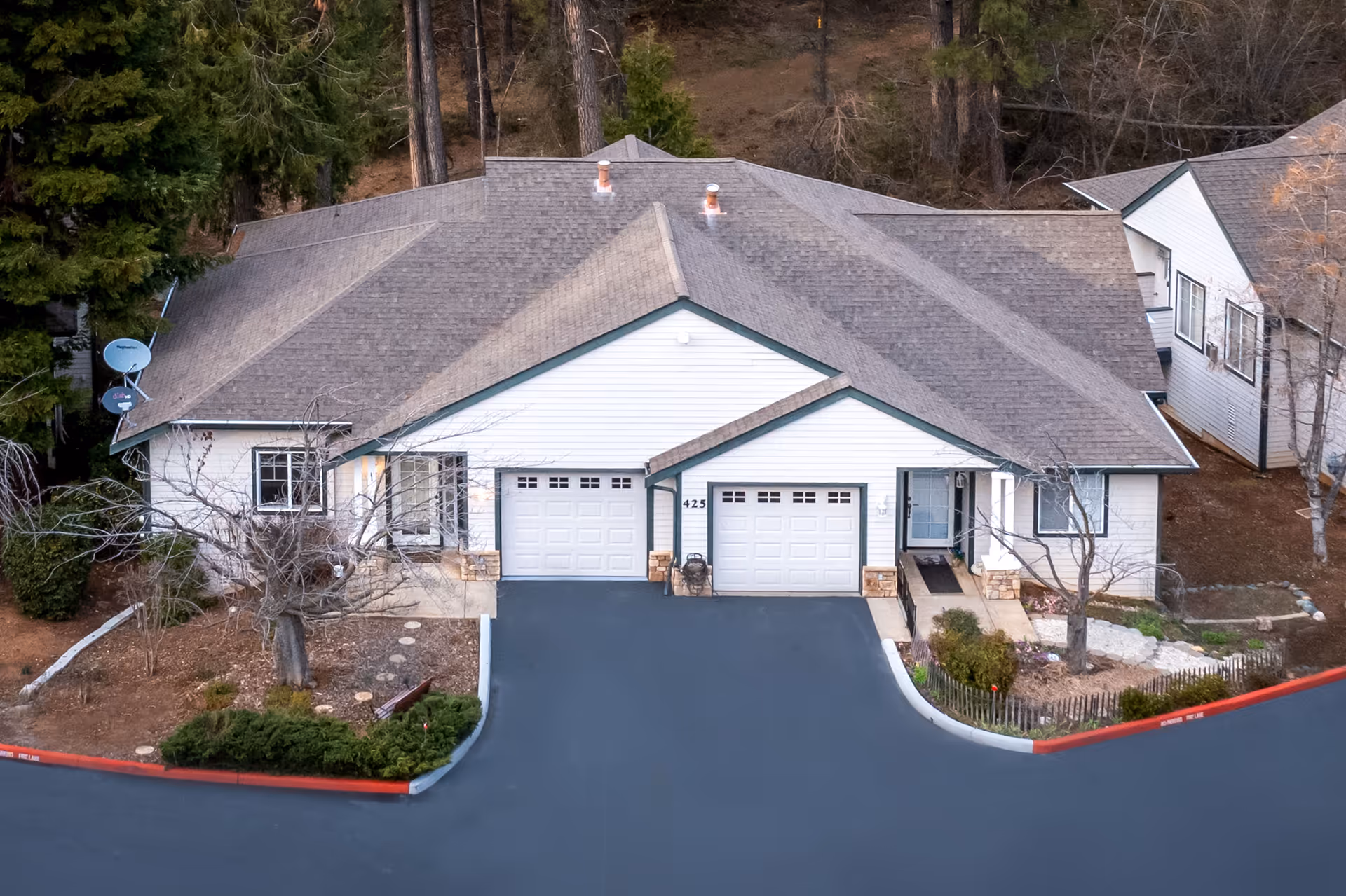 Aerial view of a single-story residential building with two attached garages, surrounded by trees and landscaping. The building has a gray shingled roof and white siding with green trim. There is a paved driveway in front and some bushes and small trees around the property.