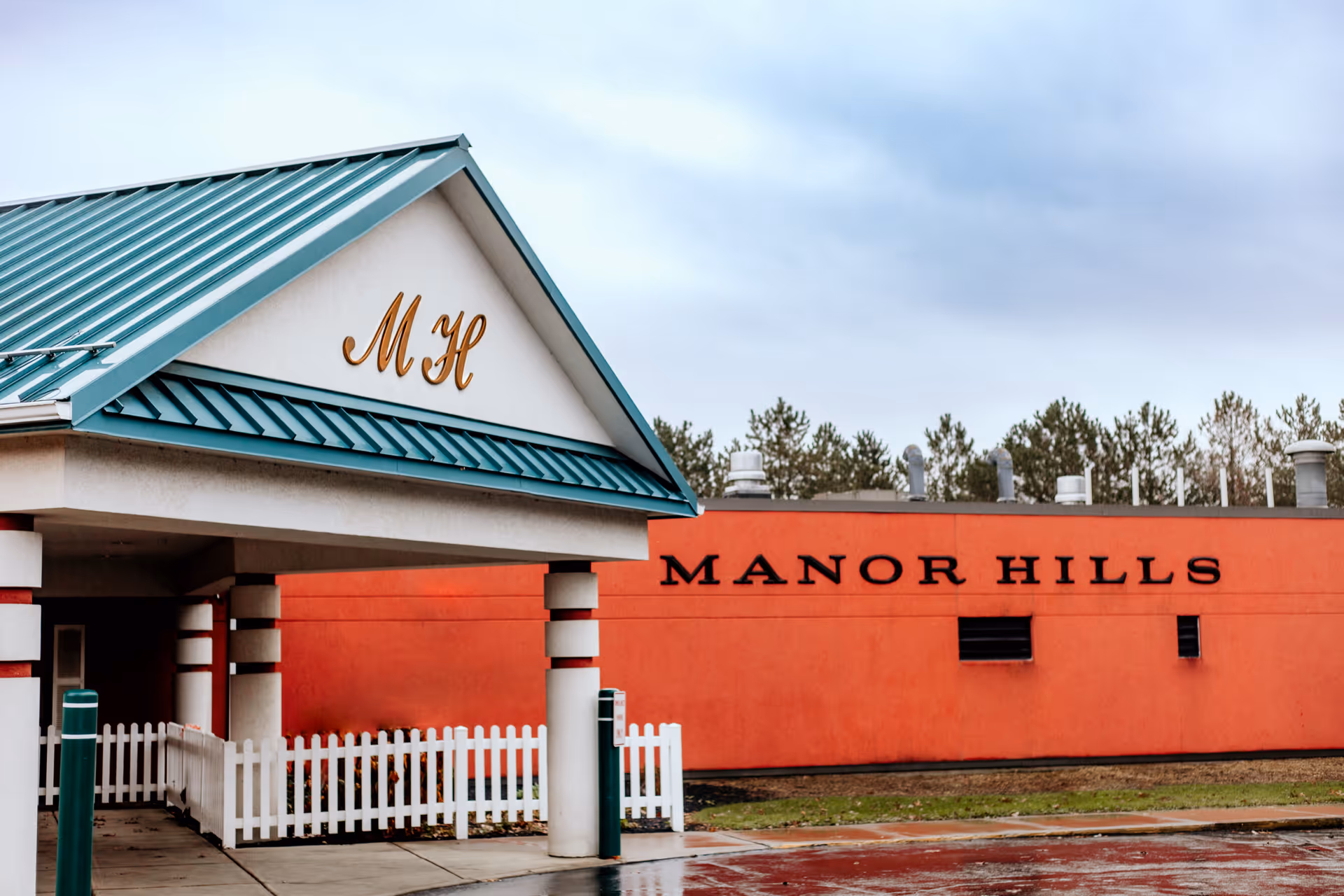Exterior view of Manor Hills senior living community building with a teal metal roof and white pillars supporting an entrance canopy. The building has a red-orange wall with the words 'MANOR HILLS' displayed prominently. There is a white picket fence near the entrance and trees in the background under a cloudy sky.