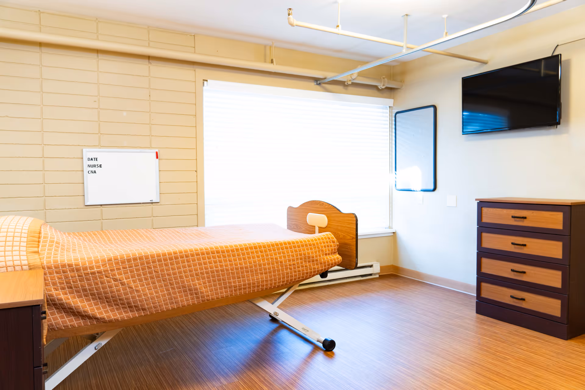 A bright senior care bedroom with a single adjustable bed, dresser, wall-mounted TV, and large window.