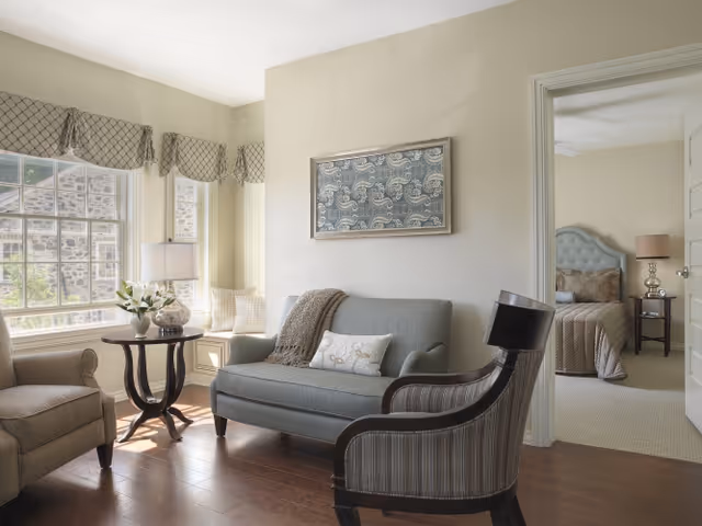 A bright sitting area with upholstered sofa and chairs, a side table by a large window, and a view into a bedroom.
