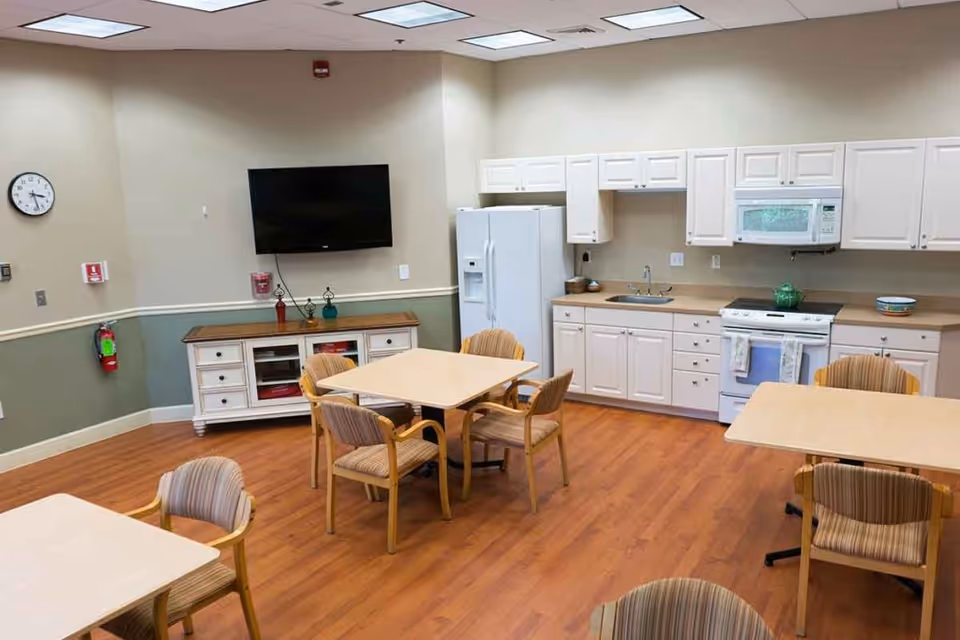 A communal kitchen and dining area with wooden floors, beige walls, and ceiling lights. The room features white cabinets, a refrigerator, stove, microwave, and sink along one wall. There are several square tables with striped cushioned chairs arranged around the room. A flat-screen TV is mounted on the wall above a white sideboard with decorative items. A clock and fire extinguisher are visible on the wall.