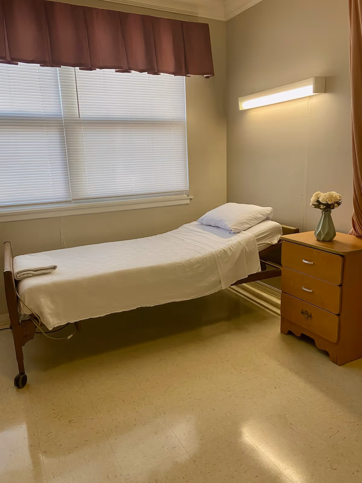 A simple room with a single hospital-style bed covered with a white sheet and pillow. Next to the bed is a wooden nightstand with three drawers and a vase with white flowers on top. The room has a large window with closed blinds and a maroon valance. A wall-mounted light fixture is above the bed.