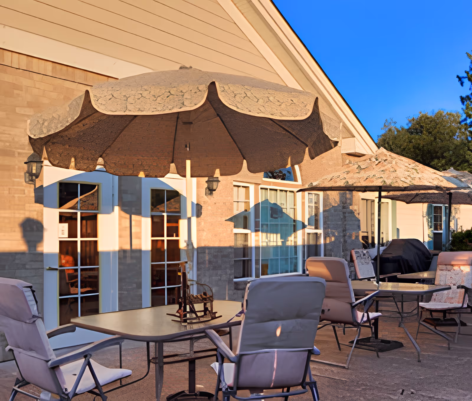 Outdoor patio area with several tables and cushioned chairs under large beige umbrellas, adjacent to a building with large windows and doors, under a clear blue sky.