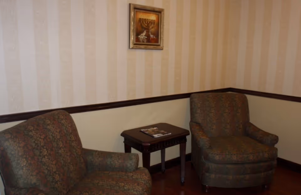 A small sitting area with two patterned armchairs and a dark wooden side table between them. A framed picture hangs on the wall above the table. The walls have a beige striped wallpaper with a dark wooden chair rail.