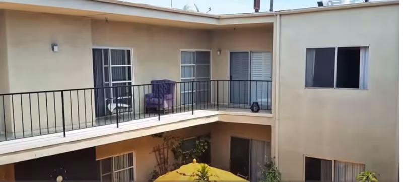 View of the exterior courtyard of West Pico Terrace Assisted Living Center showing a two-story building with beige walls, several windows and sliding doors, a black metal railing on the upper floor balcony, a purple armchair on the balcony, and a yellow umbrella over some plants in the courtyard below.
